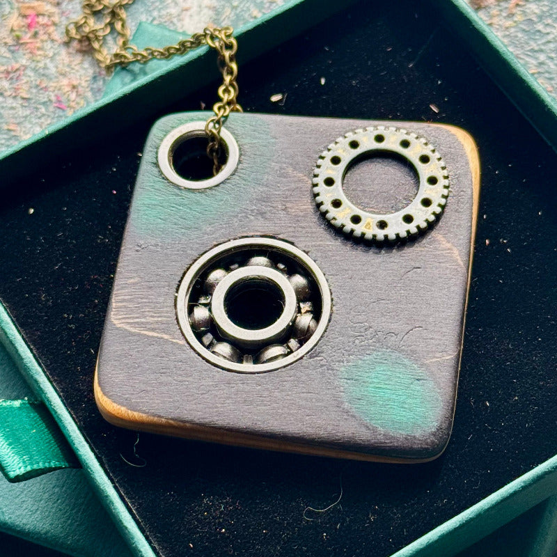 Close-up of the bearing and gear pendant in grey-green maple with an orange edge, on a brass chain.