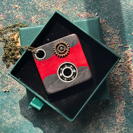 Recycled skateboard wood square pendant in red and black with three embedded metal gears and bearings, shown in a green jewellery box on a textured background.