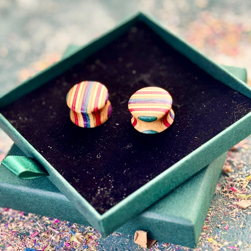 A pair of handcrafted wooden ear plugs made from recycled skateboard wood, shown in a green jewellery box. The plugs have smooth rounded faces with multi-coloured vertical colour tones including purple, teal, red, yellow, and natural maple. The background shows scattered sanding dust from the making process.