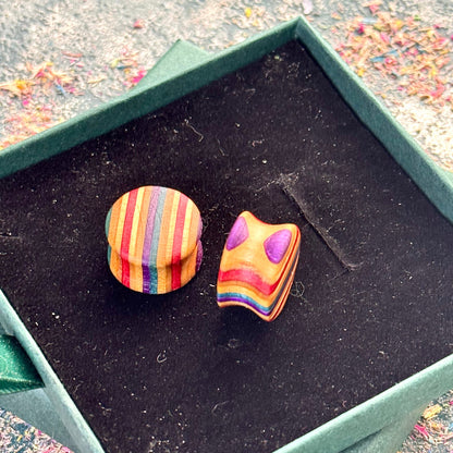 Hand-shaped wooden plugs made from recycled skateboard wood, showing bright striped colour tones in purple, red, orange and natural maple, photographed inside a green jewellery box.