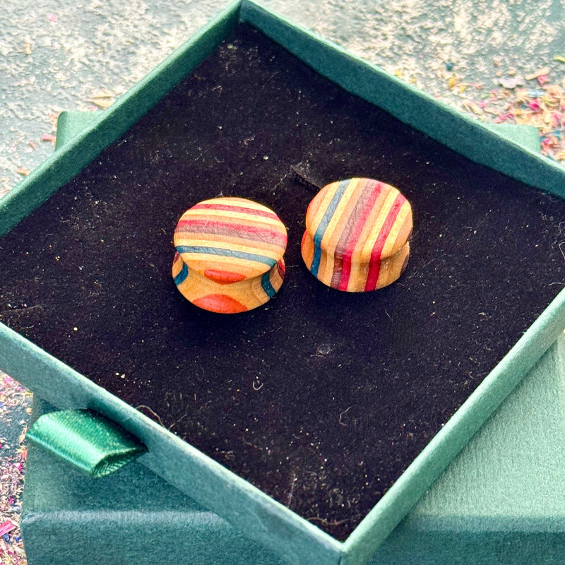 Small handcrafted wooden plugs made from recycled skateboard wood, featuring mixed colourful tones with soft rounded fronts, displayed inside a green jewellery box on a dark background.