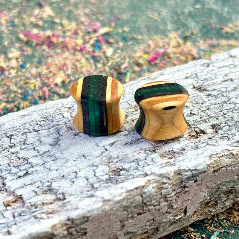 A pair of handmade wooden ear plugs shaped with a gentle hourglass curve, showing natural maple tones with deep forest-green colour accents, photographed on a weathered plank with scattered sanding dust.