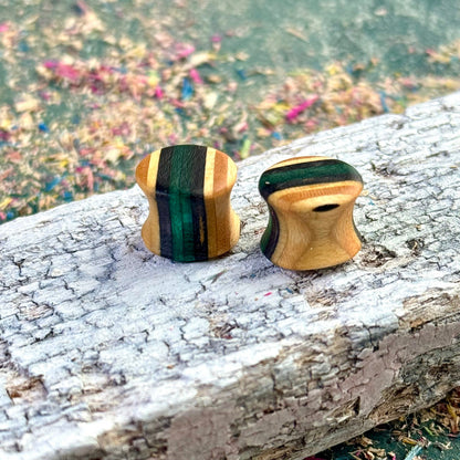 A pair of handmade wooden ear plugs shaped with a gentle hourglass curve, showing natural maple tones with deep forest-green colour accents, photographed on a weathered plank with scattered sanding dust.