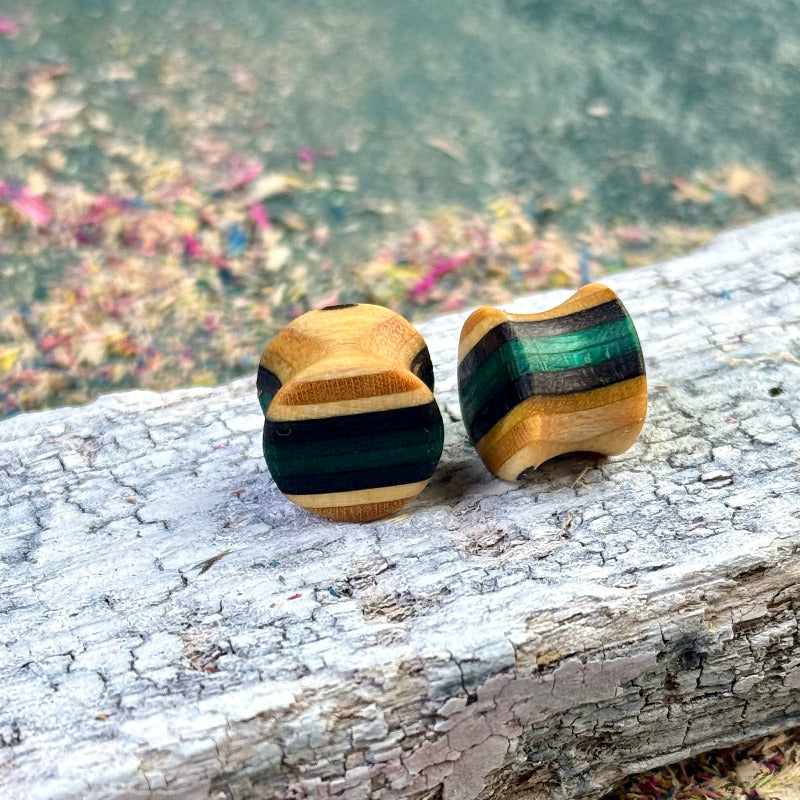 Wooden ear plugs made from recycled skateboard wood in green, black and natural colour tones, shaped with a double-flare profile and displayed on a textured wooden surface.