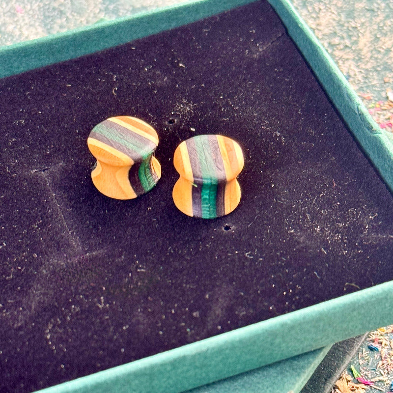 Recycled skateboard wood plugs shaped with a concave middle, showing natural maple tones with green and purple colour tones, displayed inside a green jewellery box lid on a black velvet surface.