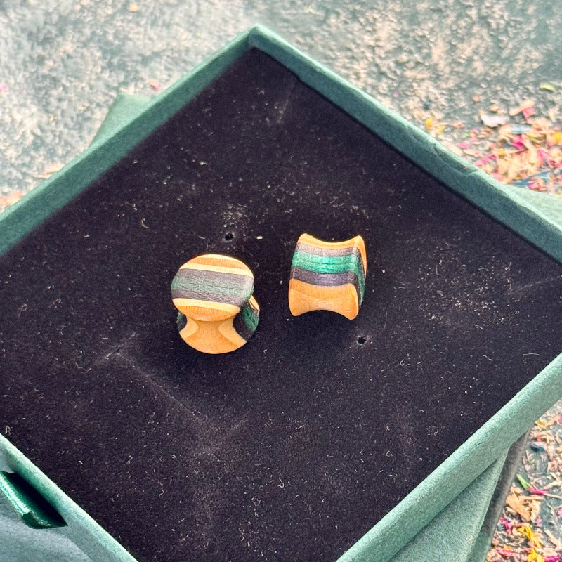Hand-carved wooden ear plugs made from recycled skateboard wood with green and natural colour tones on a dark display box