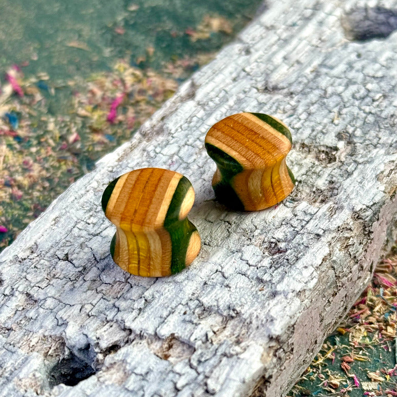 A pair of handmade recycled skateboard wood plugs in green and natural wood colour tones, shown on a textured white wood surface for close detail.