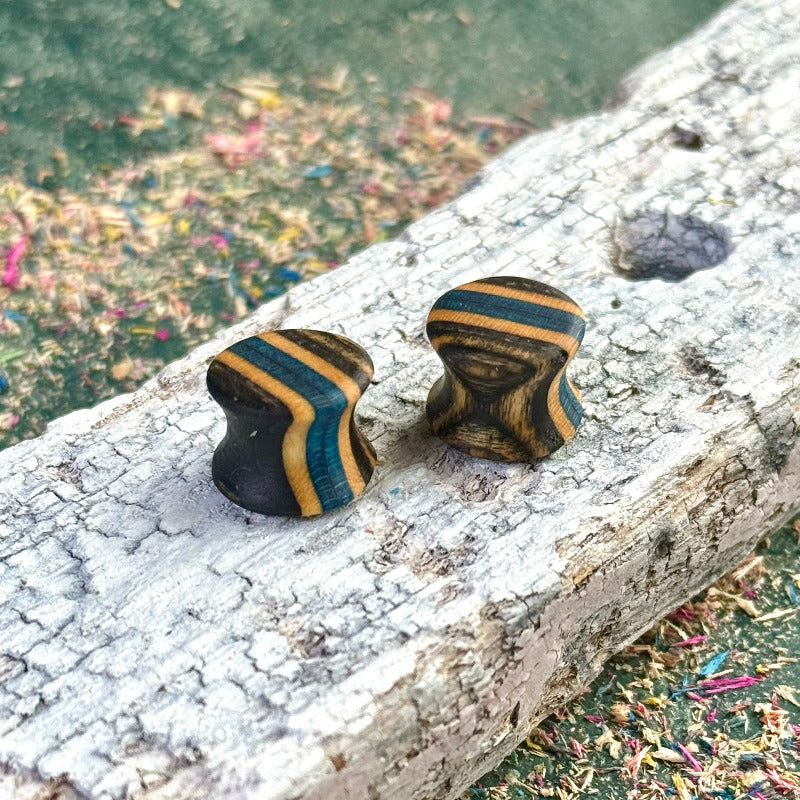 Handmade wooden plugs from recycled skateboard wood with blue, black and natural tones, shown on a textured weathered board.
