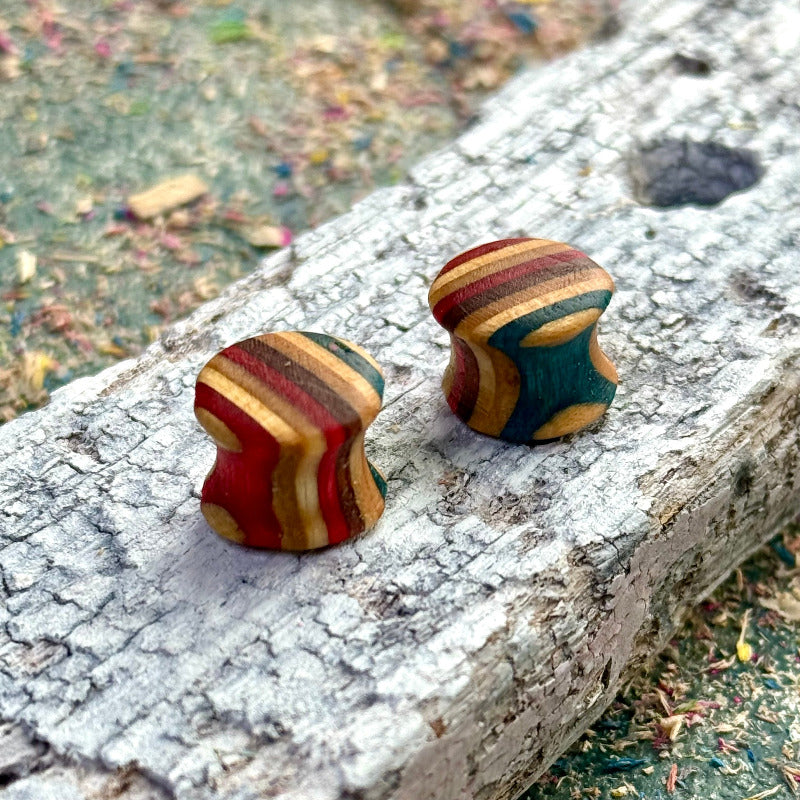 Hand-shaped wooden plugs made from recycled skateboard wood, featuring curved profiles with red, teal, mustard and natural wood colour tones, displayed on a textured piece of weathered wood outdoors.