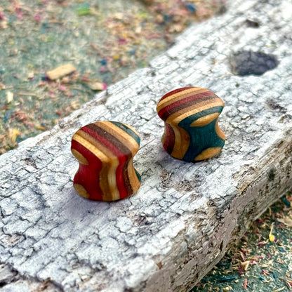 Hand-shaped wooden plugs made from recycled skateboard wood, featuring curved profiles with red, teal, mustard and natural wood colour tones, displayed on a textured piece of weathered wood outdoors.