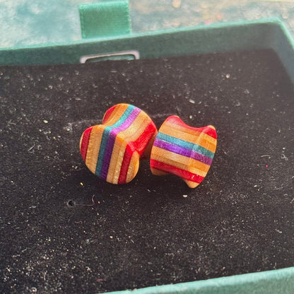 Handmade wooden ear plugs made from recycled skateboard wood with purple, red and teal colour tones, shown on a black velvet jewellery box.