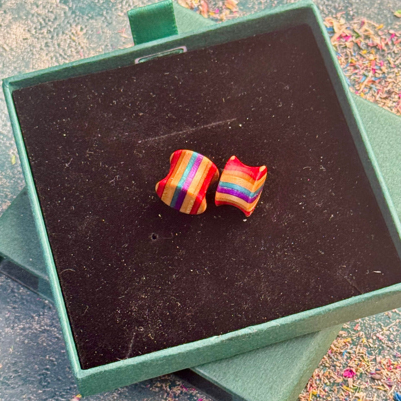 Hand-carved skateboard wood plugs with curved shape displayed in a green jewellery box, featuring red, teal, purple and rainbow colour tones.
