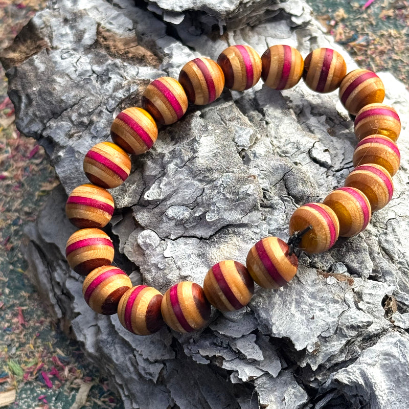 Beaded bracelet made from recycled skateboard wood in warm yellow and deep pink tones, shown on textured bark background.