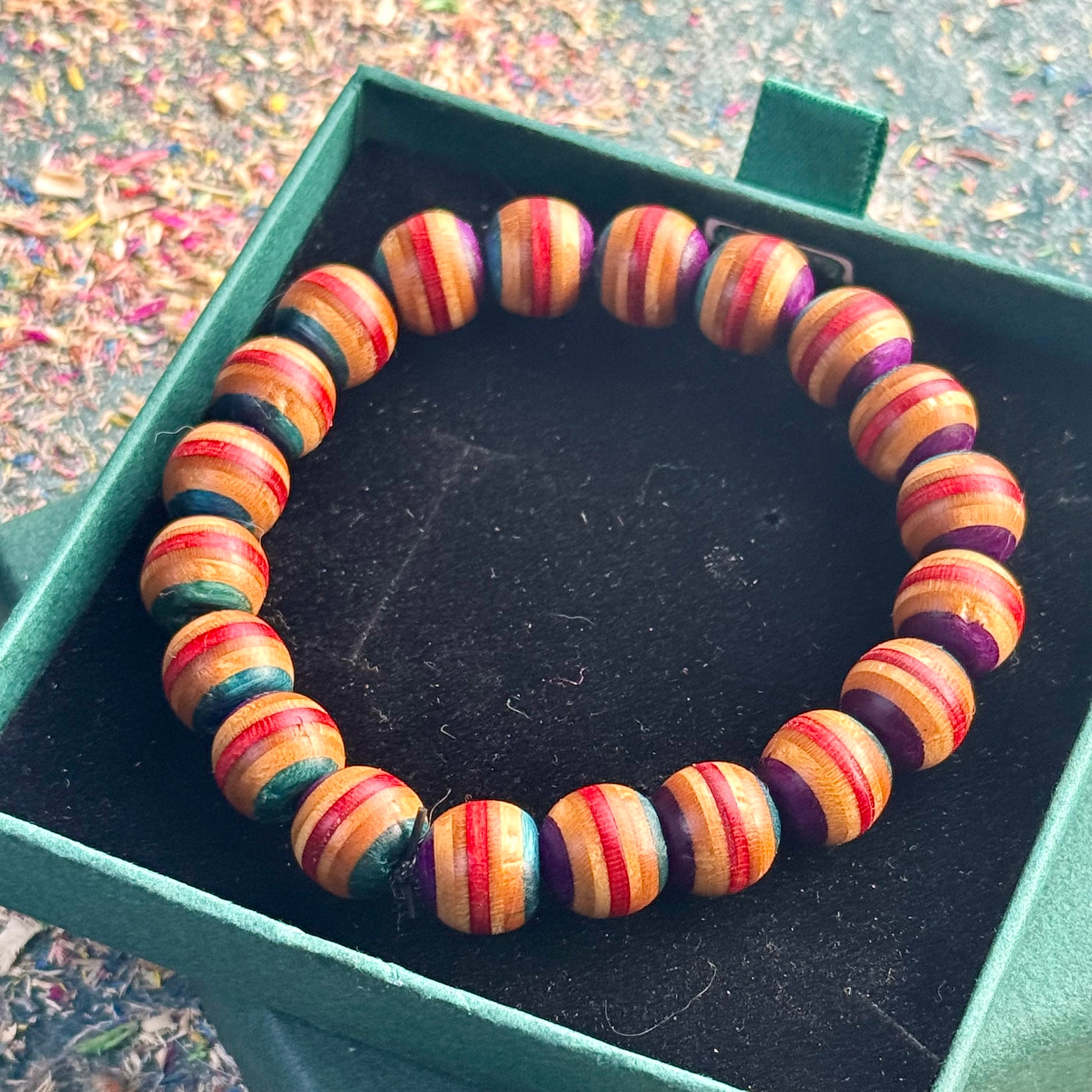 Teal, red and purple skateboard-wood beaded bracelet displayed in a green gift box on a textured background.