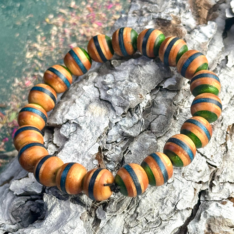 Handmade bracelet from recycled skateboard wood with round beads in natural wood tones and green and blue colour layers, shown on textured bark background.