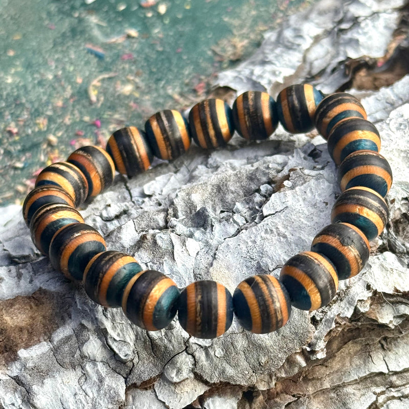 Handmade bracelet crafted from recycled skateboard wood with alternating black and brown colour tones, shown as rounded 10mm beads strung in a full circle, photographed on textured bark with a soft green background.