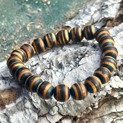 Handmade bracelet crafted from recycled skateboard wood with alternating black and brown colour tones, shown as rounded 10mm beads strung in a full circle, photographed on textured bark with a soft green background.