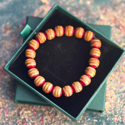 Handmade beaded bracelet crafted from recycled skateboard wood in warm yellow and red tones, displayed inside a dark velvet jewellery box with a green lid on a textured background.