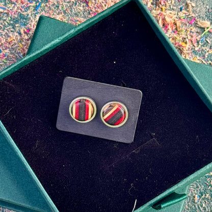 Round stud earrings crafted from recycled skateboard wood in red and black tones, displayed on a backing card inside an open green gift box.