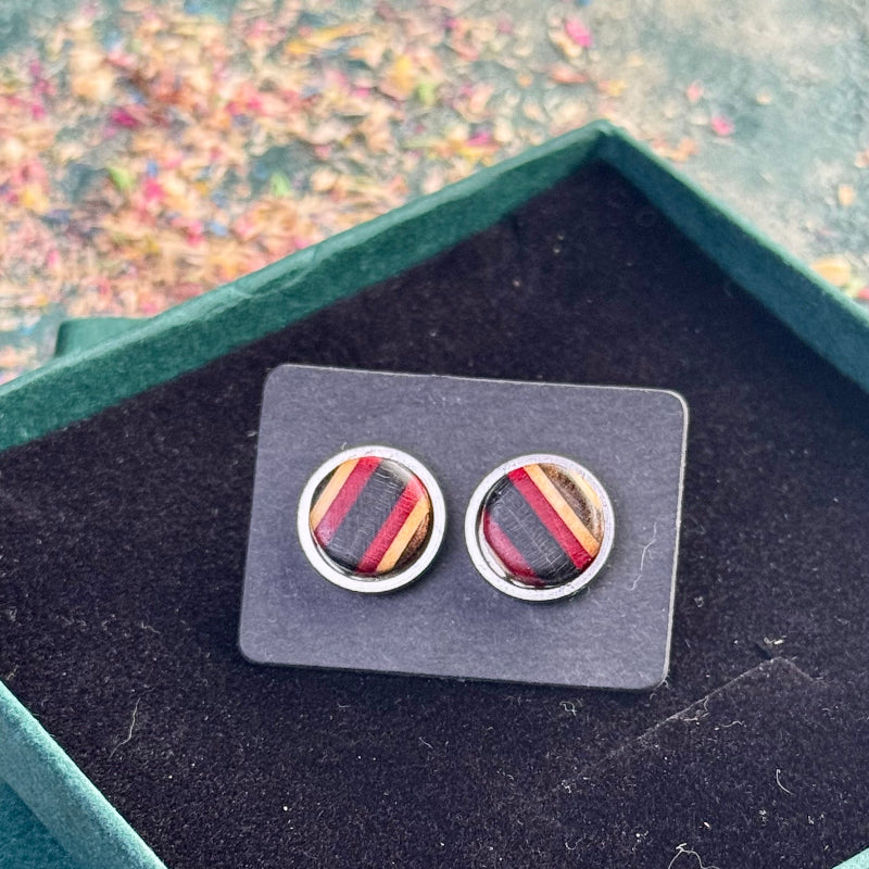 Round stud earrings made from recycled skateboard wood, showing red and black colour tones in angled stripes, photographed on a black card inside a green jewellery gift box.