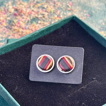 Round stud earrings made from recycled skateboard wood, showing red and black colour tones in angled stripes, photographed on a black card inside a green jewellery gift box.