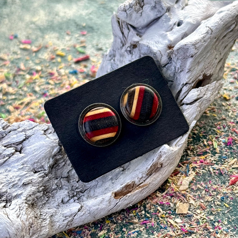 Pair of round stud earrings made from recycled skateboard wood in red and black tones, shown on a black backing card resting on textured driftwood.