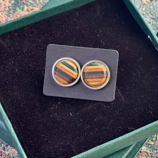 Close-up of round stud earrings made from recycled skateboard wood showing green, black and brown tones on a black card.