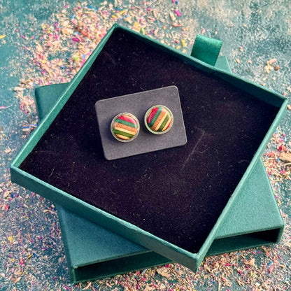 A pair of stud earrings made from recycled skateboard wood with red, green, and soft yellow tones, shown on a black card placed inside an open teal gift box on a textured workbench surface.