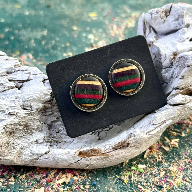 Stud earrings with pink, purple, dark green and teal striped skateboard wood shown on a dark backing card resting on driftwood.