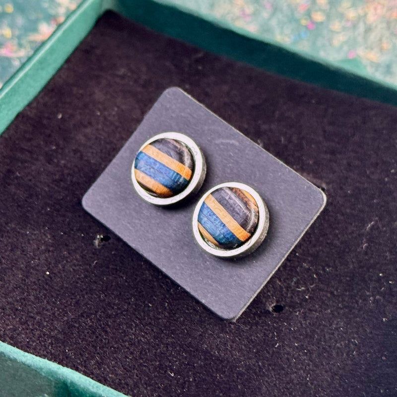 Close-up of stud earrings with blue and black striped skateboard wood resting on a dark backing card in a gift box.