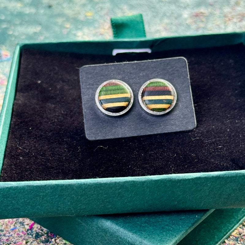 Close-up of stud earrings with green, teal and purple striped skateboard wood resting on a dark card inside a gift box.
