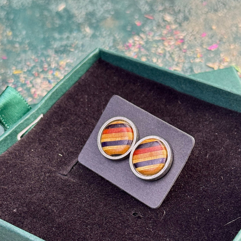Close-up of stud earrings with amber, red and purple striped skateboard wood resting on a dark display card inside a gift box