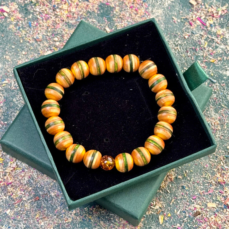 Wooden bracelet made from recycled skateboard beads in green and yellow colour tones, arranged on an elastic band and displayed in a green jewellery box, finished with one real amber bead.