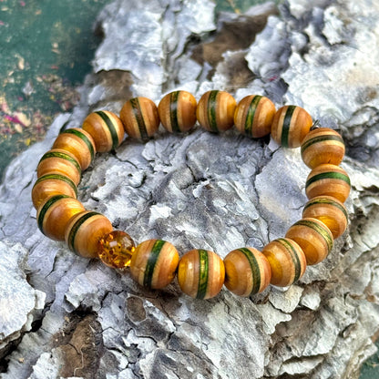 A handmade beaded bracelet crafted from recycled skateboard wood, featuring green and yellow colour tones with one genuine amber bead, displayed on textured bark outdoors.
