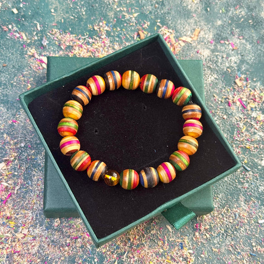 Elastic bracelet handmade from recycled skateboard wood, featuring mixed green, pink, yellow, blue, brown and natural tones, with a single real amber bead, photographed in a green presentation box on a textured background.