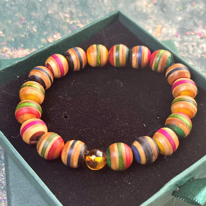 Bracelet made from recycled skateboard wood with multicoloured beads in green, yellow, pink, blue, red, and natural brown tones, finished with one real amber bead, displayed on a dark velvet box.