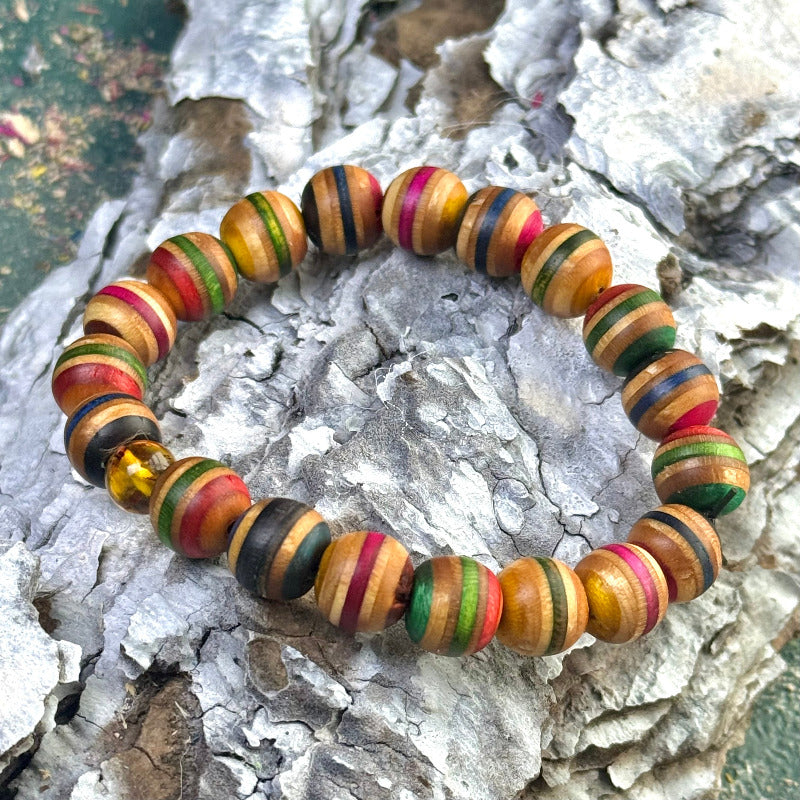 A handmade recycled skateboard bracelet featuring yellow, red, green and black colour tones. Each 10mm bead shows natural wood grain with varied stripe patterns, finished with one genuine amber bead included in the circle. The bracelet is elastic and displayed outdoors on textured bark.