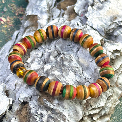 A handmade recycled skateboard bracelet featuring yellow, red, green and black colour tones. Each 10mm bead shows natural wood grain with varied stripe patterns, finished with one genuine amber bead included in the circle. The bracelet is elastic and displayed outdoors on textured bark.