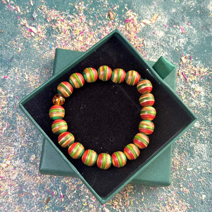 Colourful round-bead bracelet made from recycled skateboard wood in green, red and yellow tones with a single real amber bead, shown in a green gift box.