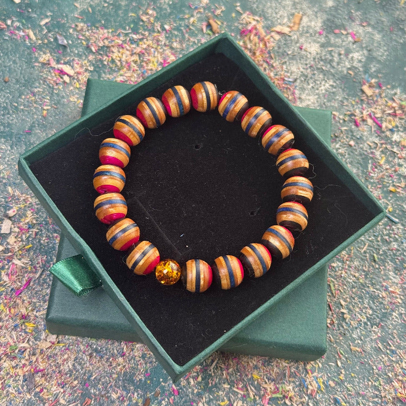 Elastic recycled skateboard bracelet featuring pink, blue and black wood tones with one real amber bead, shown in a dark green gift box.