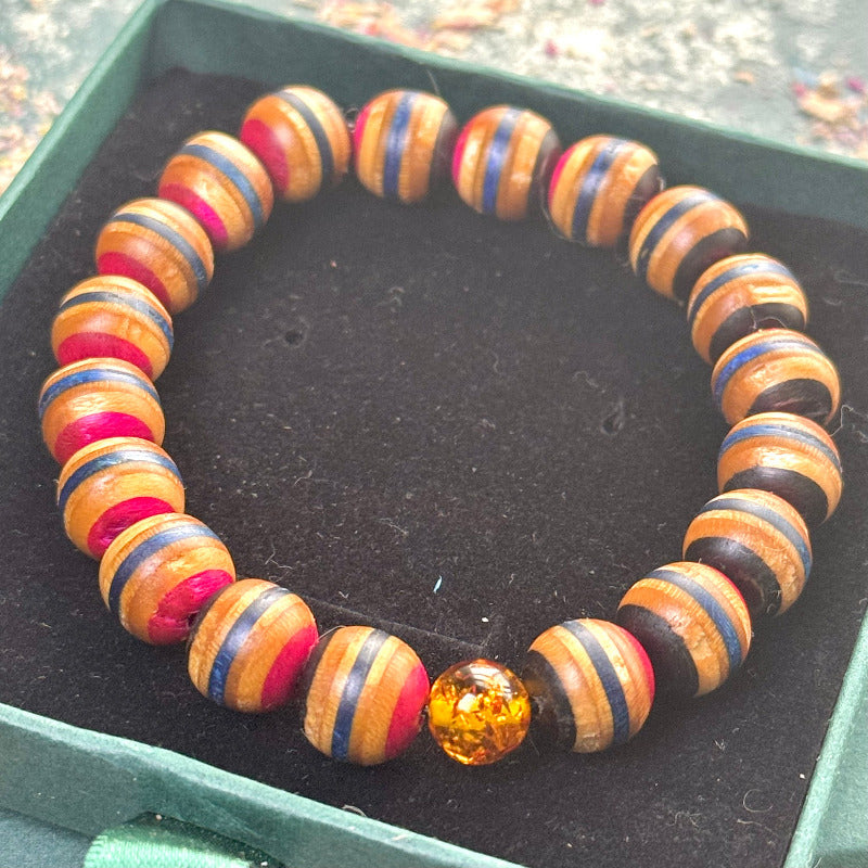Close-up of a handmade elastic bracelet made from recycled skateboard wood beads with alternating red, blue and natural brown colour tones, finished with a single genuine amber bead, displayed in a green jewellery box.
