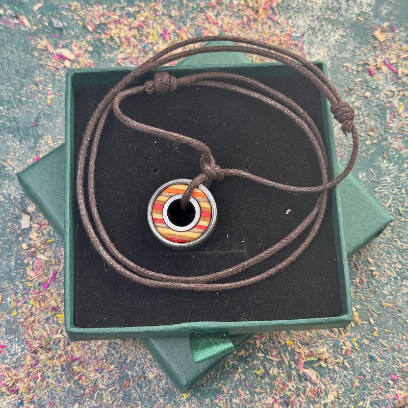 Round skateboard pendant with yellow, orange and red colour tones on a dark adjustable cord, displayed in a green gift box, photographed on a textured background.