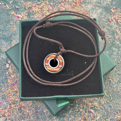 Round skateboard pendant with yellow, orange and red colour tones on a dark adjustable cord, displayed in a green gift box, photographed on a textured background.