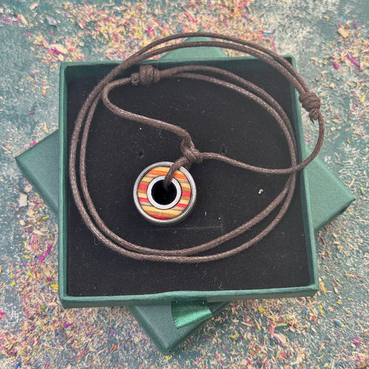 Round skateboard pendant with yellow, orange and red colour tones on a dark adjustable cord, displayed in a green gift box, photographed on a textured background.