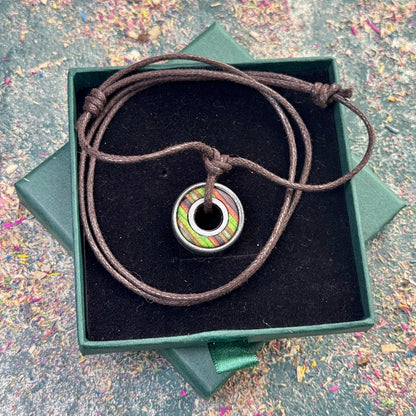 Circular recycled skateboard pendant with green, brown and purple colour tones, set inside a metal bearing and hung on a brown adjustable cord, displayed in a green gift box
