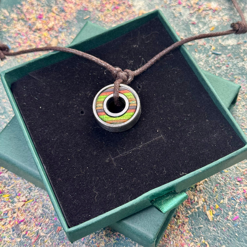 Handmade ball bearing pendant on an adjustable dark cord, featuring recycled skateboard wood in green, brown and purple colour tones, displayed in a green gift box with wood shavings in the background.