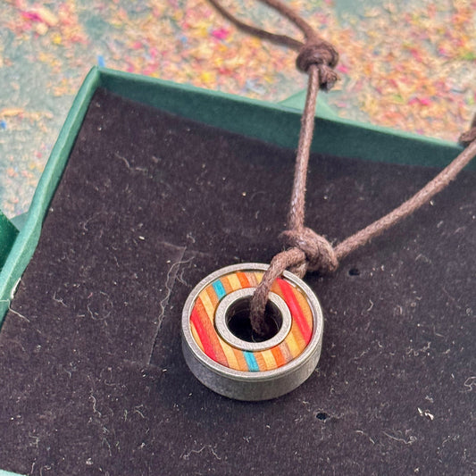 Small round pendant made from recycled skateboard wood with turquoise, red and yellow colour tones, set inside a metal bearing and hung on a brown cord