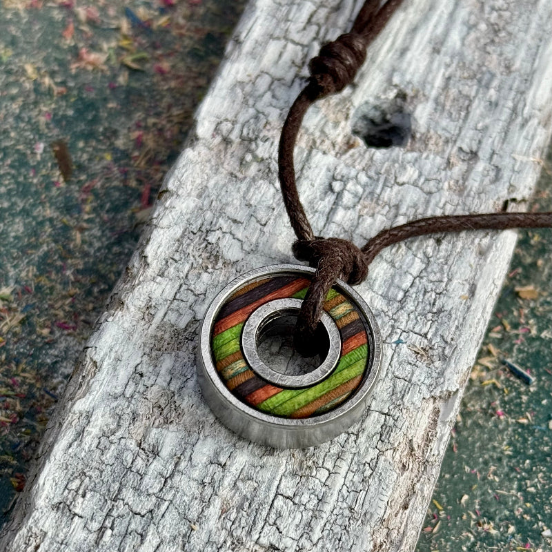 Handmade pendant with a recycled skateboard inlay featuring green, brown, and purple colour tones, set inside a steel skate bearing and hung on a brown adjustable cord, photographed on weathered wood.