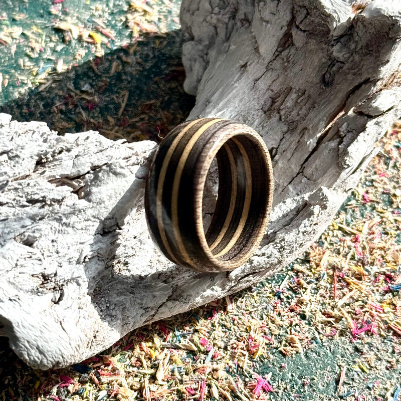 Dark grey recycled skateboard wood ring displayed on pale driftwood, showing the inner band and layered maple grain.