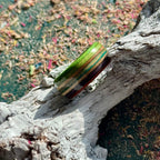 Green, purple and teal recycled skateboard wood ring resting on weathered wood, showing the full-width layered maple band.